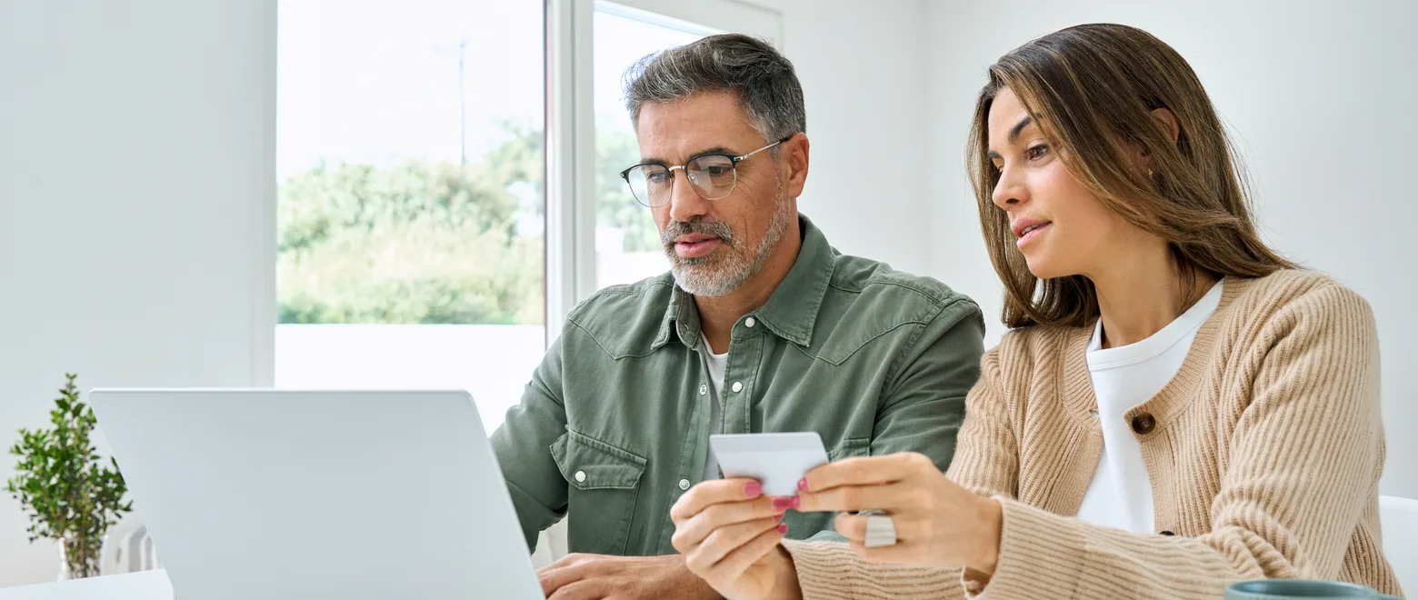 Two adults comparing plan documents and a laptop at a kitchen table