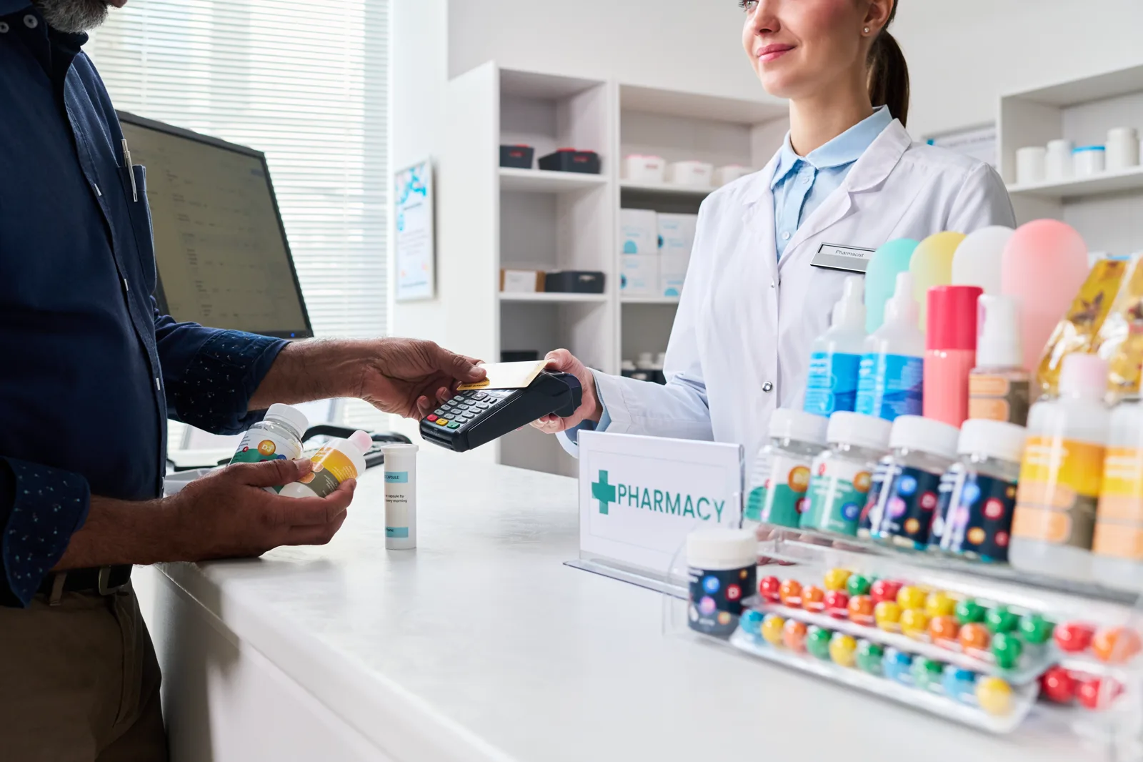 A customer paying by card at a pharmacy counter while holding prescription bottles